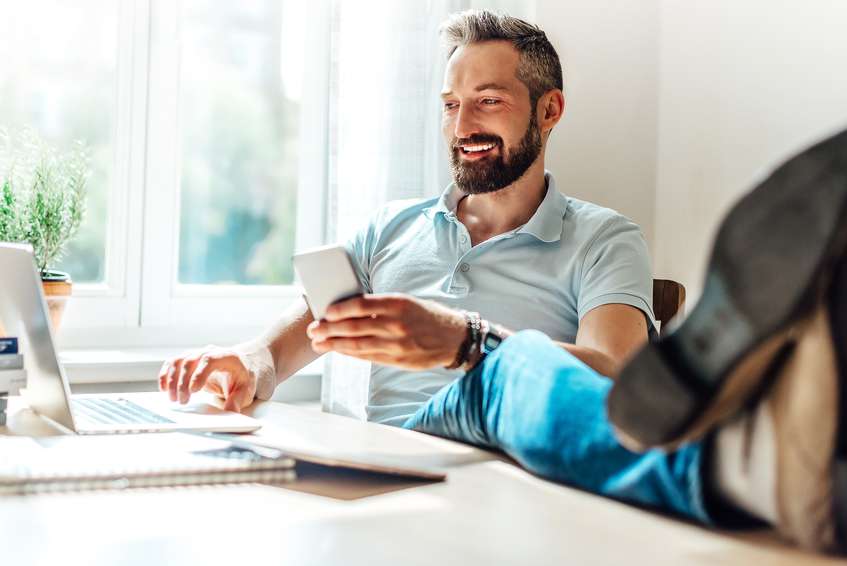 Man working from desk with his feet up, on laptop and phone smiling while working from home.