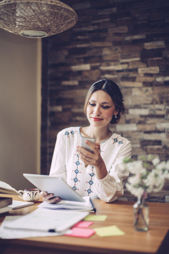 Woman sitting at dining table working from home using mobile phone and tablet.